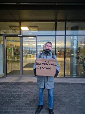 Priešais Seimo II rūmų įėjimą stovi protestuotojas su kartono dėžute, ant kurios parašyta: „Tik surenku čekius ir į Seimą“